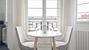 Une pièce avec une table ronde et deux chaises, un radiateur et une fenêtre donnant sur des immeubles.