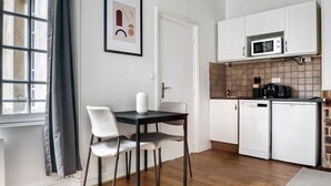 A compact kitchen with white cabinets, a microwave, and a toaster. There is a dining area with a table and chairs, and a framed picture on the wall. - Blueground | Le Marais, nr Les Halles (Paris)