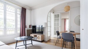 Un saló modern amb un sofà, una taula de centre, un televisor sobre un moble de fusta, una taula de menjador amb cadires, i finestres grans amb cortines.