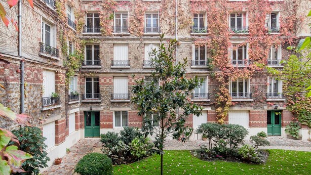 Un edifici de diverses plantes amb un pati interior, amb balcons i baranes, una porta verda i una petita zona de jardí amb plantes i un arbre.