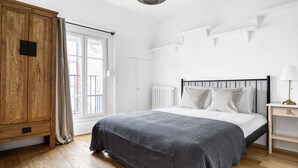 A bedroom with a wooden wardrobe, a bed with white bedding, a radiator, and a bedside table with a lamp. - Blueground | Ecole Militaire, furnished, balcony (Paris)