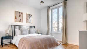 Ein Schlafzimmer mit einem Bett, zwei Nachttischen, einem Fenster mit Vorhängen und zwei abstrakten Bildern an der Wand.