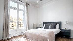 Un dormitorio con un ventanal grande, una cama con cubrecama blanco, dos mesitas de noche con lámparas y un radiador.