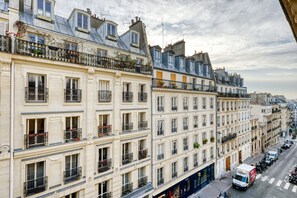 Uma fileira de edifícios parisienses clássicos, com varandas e janelas uniformes, uma rua com carros estacionados e um céu claro.