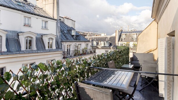 A rooftop terrace with a table and chairs, surrounded by lush greenery and overlooking a cityscape with traditional European architecture. - Blueground | Palais Royal, balcony, nr Louvre (Paris)