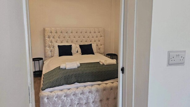Une chambre à coucher avec une tête de lit capitonnée, un lit recouvert d’une couverture verte, deux tables de chevet noires et une prise de courant blanche sur le mur.