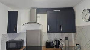 Une cuisine de taille réduite, avec des meubles de couleur sombre, un four à micro-ondes, un grille-pain, une bouilloire et une horloge murale.