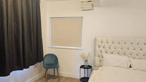 Une chambre avec un lit, une fenêtre avec des stores, une chaise, une petite table avec un vase et un climatiseur fixé au mur.