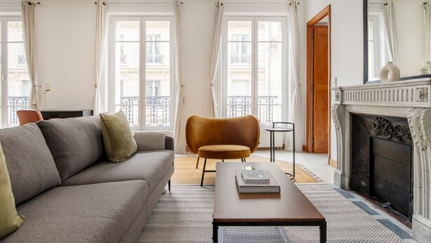 Un salon comprenant un canapé, un fauteuil et une table basse. On y trouve une cheminée et, grâce à de grandes fenêtres, une vue sur les bâtiments environnants.