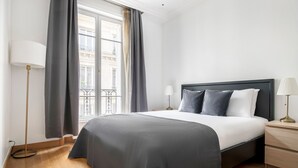 Une chambre d’hôtel moderne dotée d’un grand lit, de deux tables de chevet, d’une lampe sur pied et offrant une vue sur la ville grâce à de grandes fenêtres.