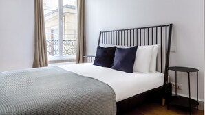 A neatly arranged bedroom with a bed, a bedside table, and a window with curtains. - Blueground | Arc de Triomphe, nr Champs Elysées (Paris)