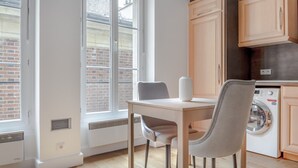 A modern kitchen with wooden cabinets, a built-in washing machine, and a dining area with a table and chairs. - Blueground | Arc de Triomphe, nr Champs Elysées (Paris)