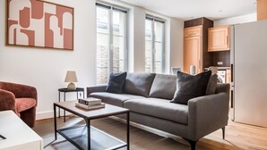A modern living room with a grey sofa, a coffee table, and a red armchair. There is a window with blinds, a lamp, and a bookshelf with books. - Blueground | Arc de Triomphe, nr Champs Elysées (Paris)