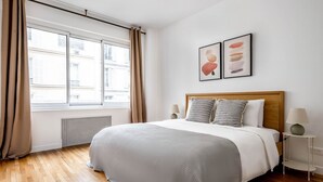 A bedroom with a large bed, a window with curtains, a radiator, a bedside table with a lamp, and two framed artworks on the wall. - Blueground | Miromesnil, furnished, elevator (Paris)