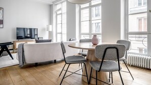 A modern living room with a round dining table, two chairs, a sofa, a TV, and large windows.