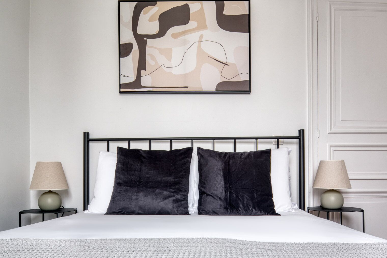 A neatly made bed with a black headboard, two bedside lamps, and a large abstract painting above it.