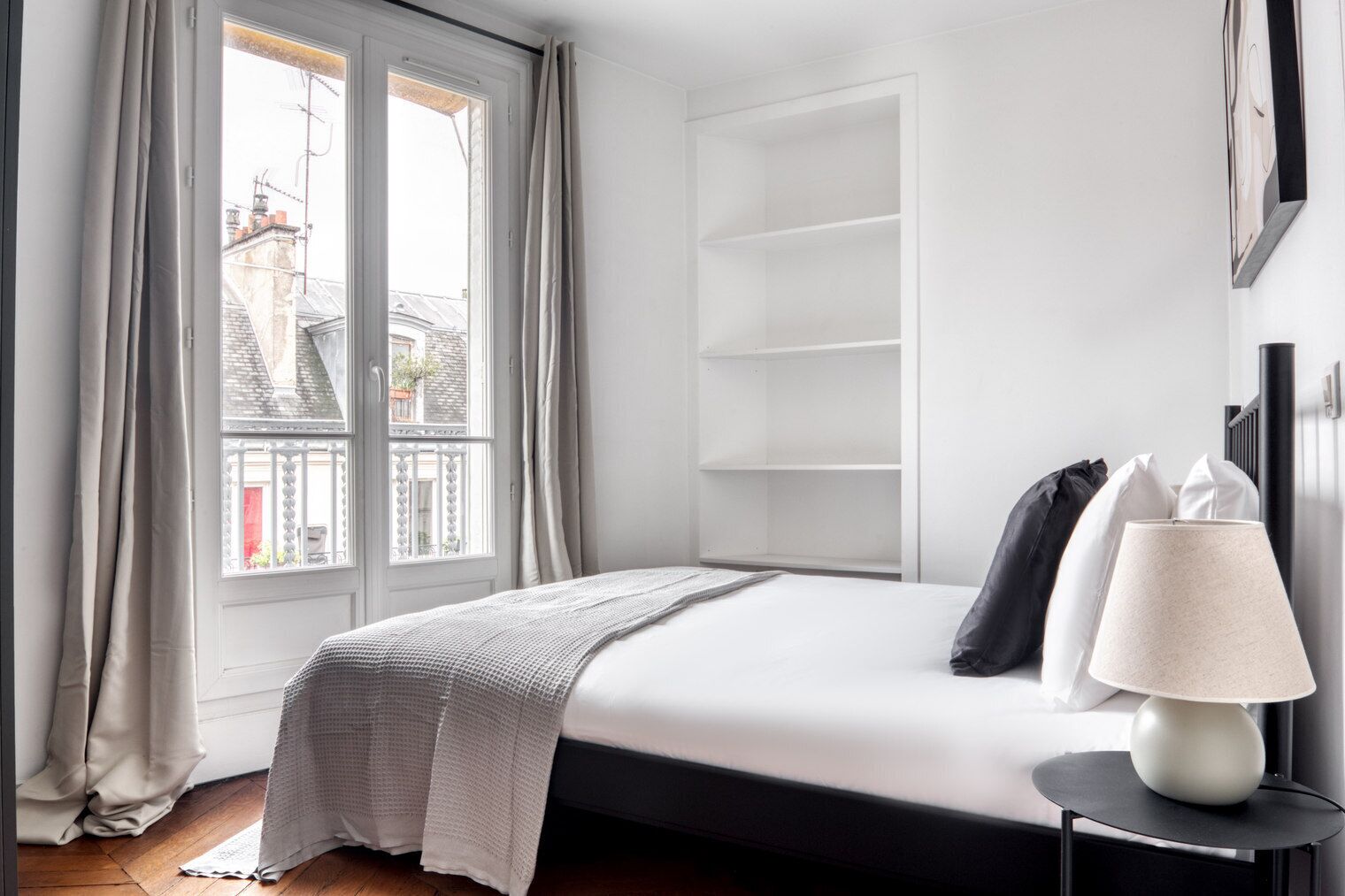 Una camera da letto moderna con un'ampia finestra, un letto con copripiumino bianco, un comodino con lampada e una libreria a muro.