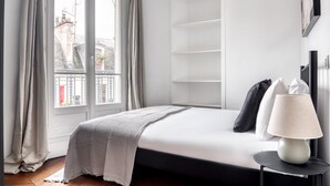 Ein modernes Schlafzimmer mit großem Fenster, einem Bett mit weißer Bettdecke, einem Nachttisch mit Lampe und einer integrierten Regaleinheit.