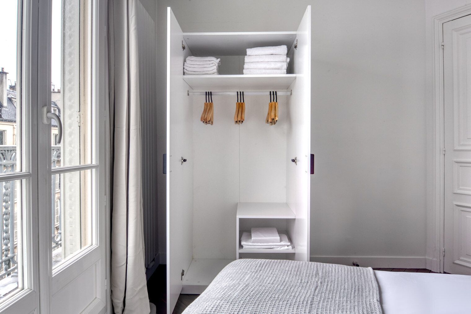 Une chambre avec un lit, une armoire blanche où sont rangées des serviettes, et une vue sur les bâtiments par la fenêtre.