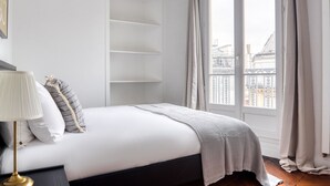 Ein Schlafzimmer mit Bett, Nachttisch, Lampe, Fenster mit Vorhängen und Blick auf Gebäude.