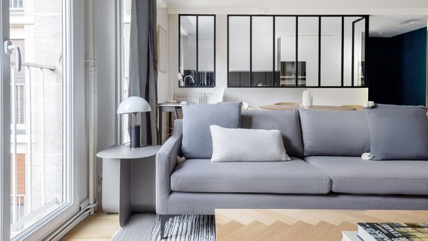 A modern living room with a grey sofa, a side table with a lamp, and a view of the kitchen through a glass door. - Blueground | Porte de Champerret, elevator (Paris)