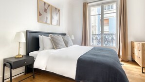 A neatly arranged bedroom with a bed, bedside table, and a view of the outside balcony. - Blueground | Rochechouart, nr Rue de Martyrs (Paris)