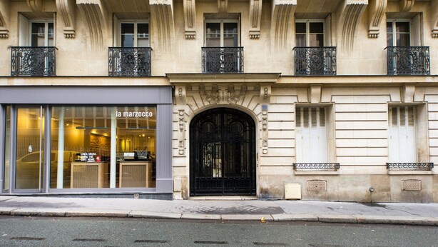 A building with a large glass storefront labeled "la marzocco" and a black ornate entrance door. - Blueground | Rochechouart, nr Rue de Martyrs (Paris)