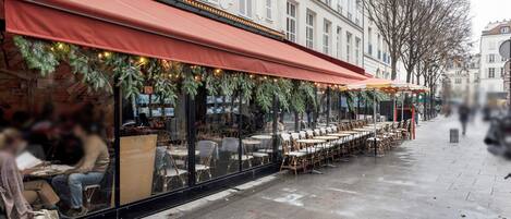 An outdoor café with tables and chairs under a red awning, decorated with greenery and lights.