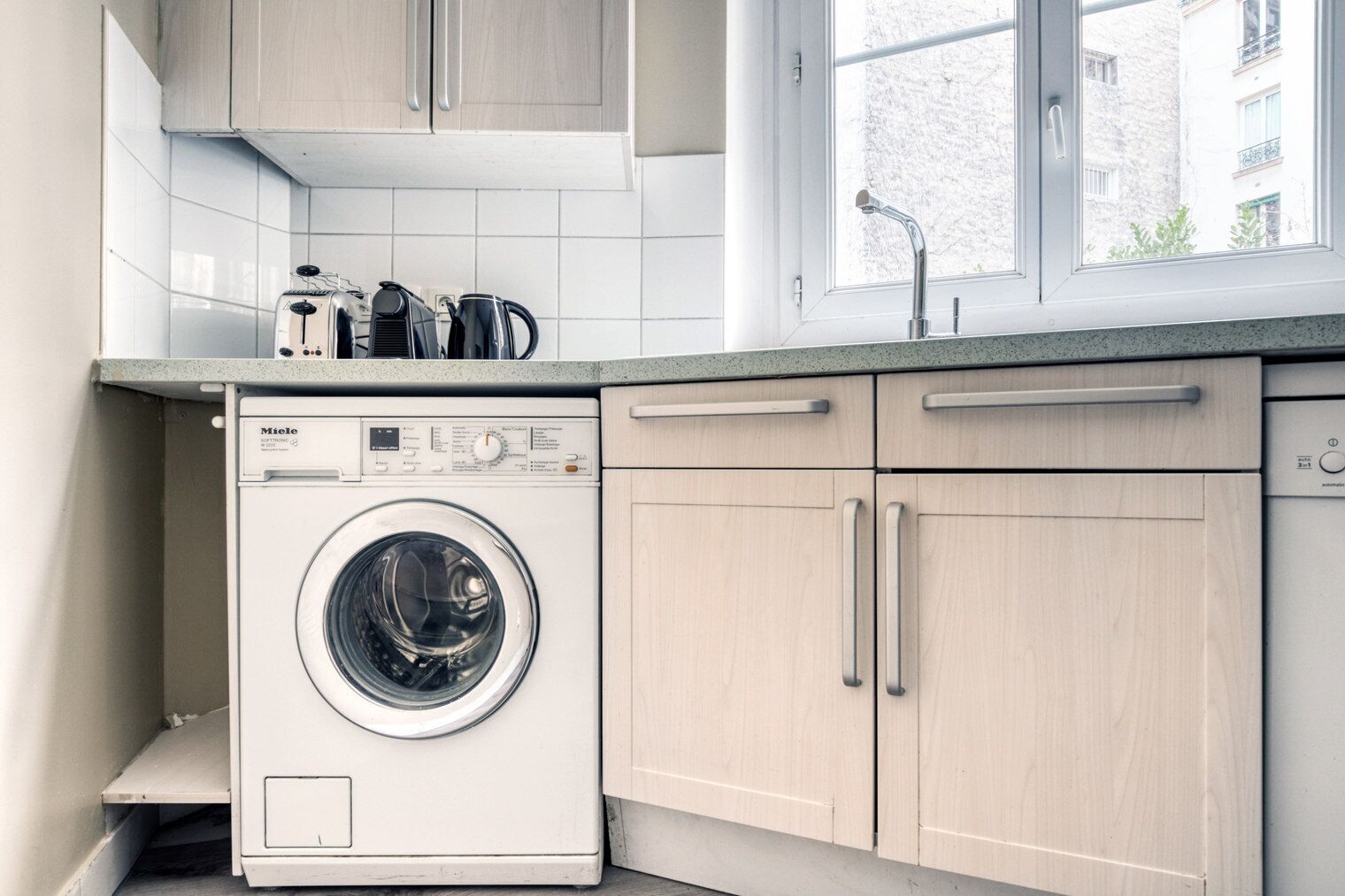 A compact kitchen with a front-loading washing machine, wooden cabinets, and a window with a view of buildings.