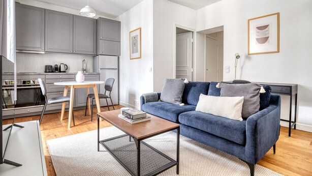 Un salon moderne avec un canapé bleu, une table basse et un coin cuisine doté de meubles gris et d’une table à manger.