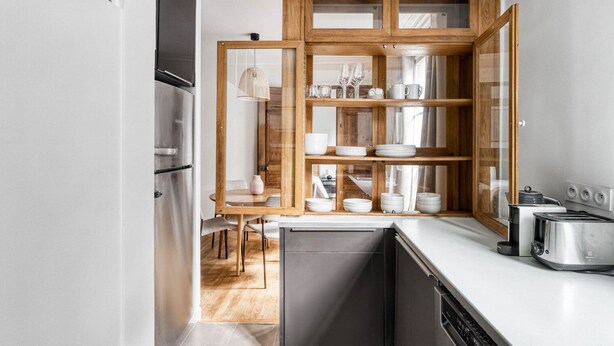 A compact kitchen with a built-in oven, a dishwasher, and a wooden cabinet with glass doors displaying white dishes and glasses.