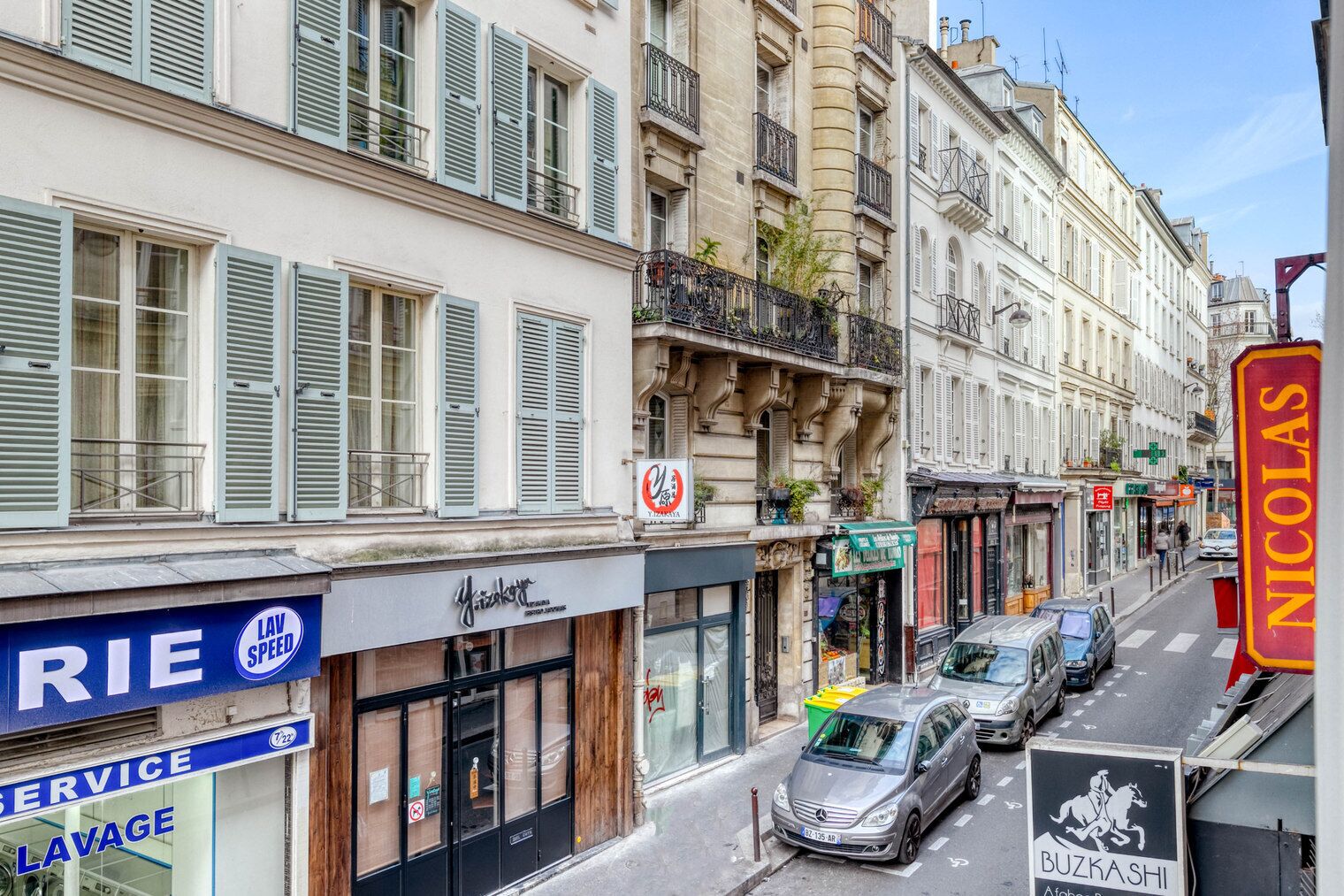 A street view with buildings, a sign for 'NICOLAS', and parked cars.