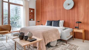 A modern bedroom with a large bed, wooden panel wall, bedside tables, a desk with books and vases, and a view of greenery outside. - Blueground | Saint Georges, terrace, nr Montmartre (Paris)
