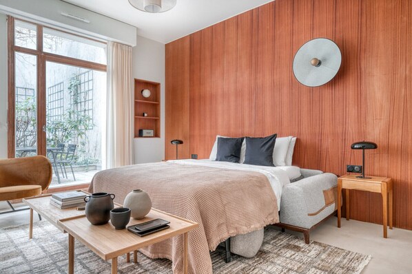 A modern bedroom with a large bed, wooden panel wall, bedside tables, a desk with books and vases, and a view of greenery outside. - Blueground | Saint Georges, terrace, nr Montmartre (Paris)
