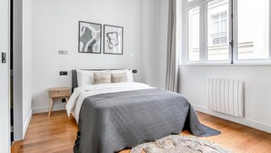 Un dormitorio con cama, mesita de noche, ventana con cortinas y dos cuadros abstractos colgados en la pared.