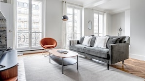 Una sala de estar moderna con un sofá gris, una mesa de centro redonda y una butaca naranja. Ventanas amplias con cortinas blancas ofrecen luz natural y vistas a edificios.