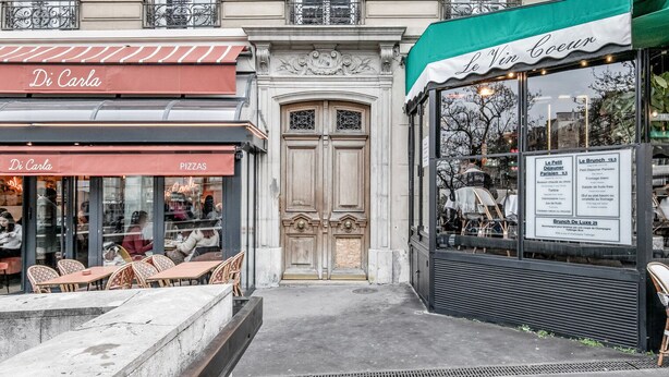 Vista de la calle con una cafetería llamada 'Di Carla' que ofrece pizzas, y otra cafetería 'Le Vin Coeur' con terraza.