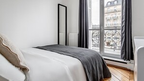Un dormitorio minimalista con un ventanal grande, una cama con ropa de cama blanca y vista a un edificio en la calle.