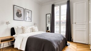 A bedroom with a bed, a bedside table, a window with curtains, and a door. - Blueground | Raspail/Rennes, balcony, furnished (Paris)