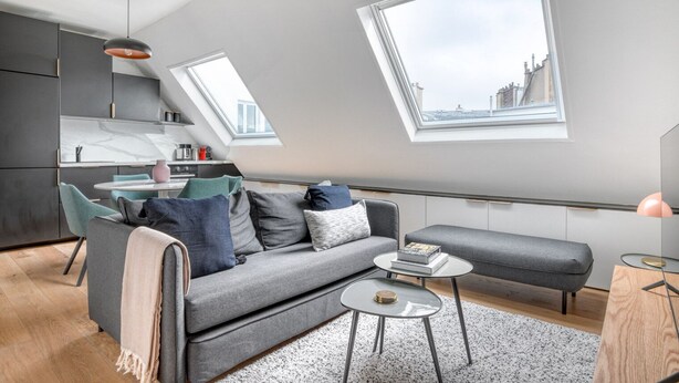 Moderni loft-tyylinen asunto, jossa on harmaa sohva, pieni pyöreä sohvapöytä sekä ruokailutila, jossa on valkoinen pöytä ja tuolit.