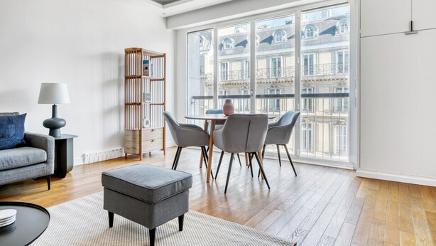 A modern living room with a grey sofa, a small table with a lamp, a grey ottoman, and a dining area with four chairs.