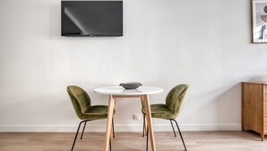 Ein schlicht eingerichteter Raum mit einem runden Tisch, zwei grünen Stühlen, einem an der Wand montierten Fernseher und einem Holzschrank.