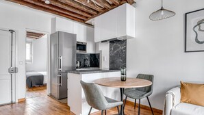 A modern kitchen with white cabinets, a marble backsplash, and a built-in oven. There is a dining area with a round table and two chairs. - Blueground | Sèvres, near shopping (Paris)