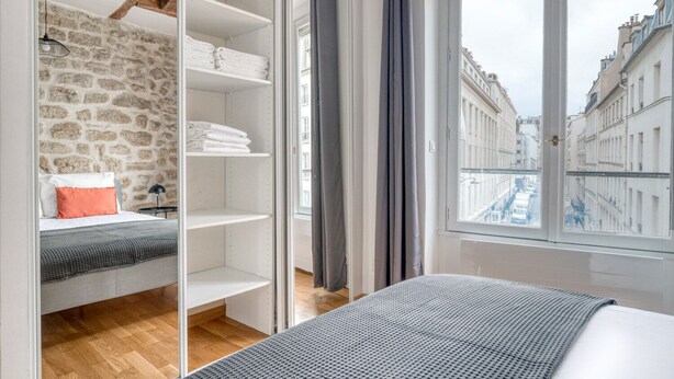 A bedroom with a stone wall, a bed with a grey blanket, a wardrobe with shelves, and a view of buildings through a window. - Blueground | Sèvres, near shopping (Paris)