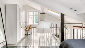Tilava ullakkohuone, jossa on korkea katto, työpöytä tuolilla, patteri ja portaikko, joka johtaa ylempään kerrokseen.