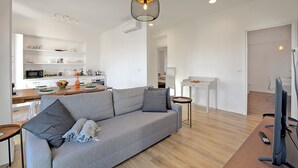 A modern living room with a grey sofa, a wooden coffee table, and a TV on a wooden stand. There is a kitchen in the background with white cabinets and a dining area. - Luce - Lake View Balcony Apartment in Stresa Hill, Stresa, Italy (Stresa)