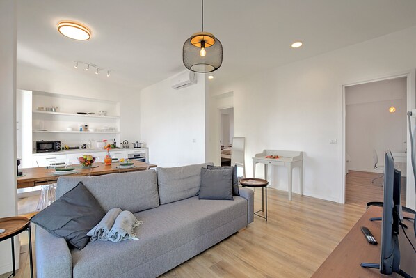 A modern living room with a grey sofa, a wooden coffee table, and a TV on a wooden stand. There is a kitchen in the background with white cabinets and a dining area.