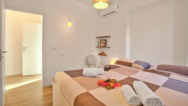 A hotel room with a large bed, a hanging light fixture, a wall-mounted air conditioner, and a door leading to another room. - Luce - Lake View Balcony Apartment in Stresa Hill, Stresa, Italy (Stresa)