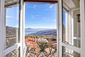 A balcony with a table and chairs, overlooking a scenic landscape with hills and a body of water. - Luce - Lake View Balcony Apartment in Stresa Hill, Stresa, Italy (Stresa)