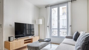 Un salon moderne avec un téléviseur à écran plat posé sur un meuble en bois, un pouf gris, un canapé gris et une grande fenêtre donnant sur des immeubles.